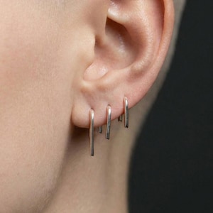 Peut inclure: Trois boucles d'oreilles en forme de U de couleur argentée sont insérées dans le lobe de l'oreille. Chaque boucle présente une finition métallique lisse et un design en barre qui pend verticalement, créant un aspect propre et uniforme.