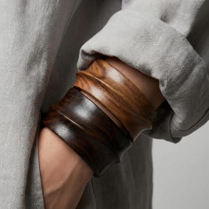 May include: Two wooden bracelets, one dark brown and one light brown, are stacked on a wrist. The bracelets have a curved, textured design. The person is wearing a gray long-sleeved shirt.