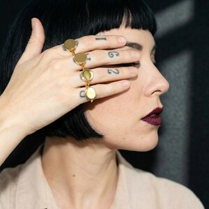 side profile with bob  hairstyle and deep lip color, hand covering the eyes layered with multiple gold signet rings, varied finishes catching light across the fingers, small tattoos adding rhythm, soft fabric backdrop with a warm, intimate feel - Rannka