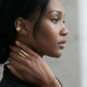 Modern sculptural gold ring worn on the hand as the focal point, styled alongside a minimalist gold ear cuff and a wide cuff bracelet, shown on a model in side profile with clean lines, soft natural light, and a refined editorial setting emphasizing contemporary jewelry styling.