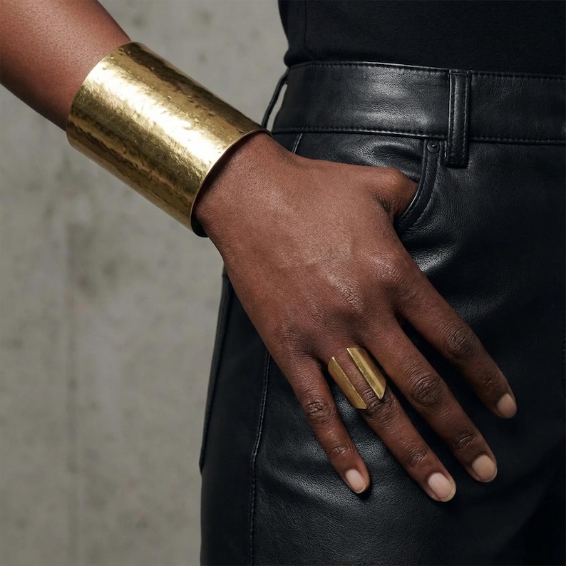 Modern sculptural gold ring worn on the hand as the primary focus, paired with a wide hammered gold cuff bracelet, styled against black leather trousers in a clean editorial setting with muted industrial tones.