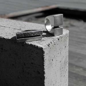 Two silver sculptural rings resting on a textured concrete block — one with a bold open-square design, the other a split hammered bar form. Industrial and architectural in style, reflecting Rannka’s minimalist handcrafted jewelry aesthetic.