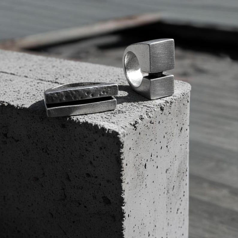 Two silver sculptural rings resting on a textured concrete block — one with a bold open-square design, the other a split hammered bar form. Industrial and architectural in style, reflecting Rannka’s minimalist handcrafted jewelry aesthetic.