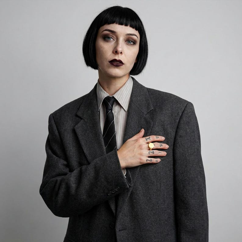 portrait with bob and dark lip, oversized tailored suit with shirt and tie, hand placed on chest showing gold signet ring, subtle finger tattoos, clean background with soft, even light and a restrained, composed mood