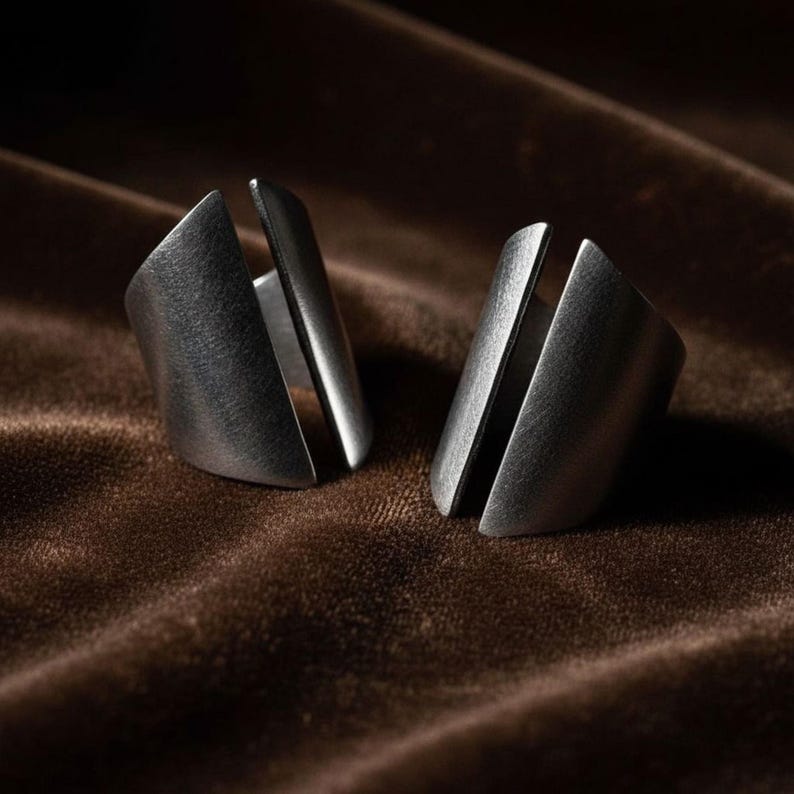 Two silver open-ended statement rings displayed on soft brown velvet fabric, showcasing their sculptural geometric shape and brushed metal finish under warm light.