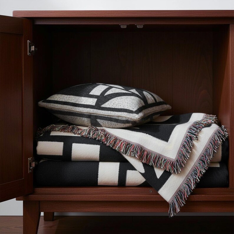 Folded black and cream geometric woven blanket with multicolor fringed edges stored neatly in a wooden cabinet, paired with a matching patterned pillow. The clean lines and graphic design bring a structured, contemporary touch to the space.