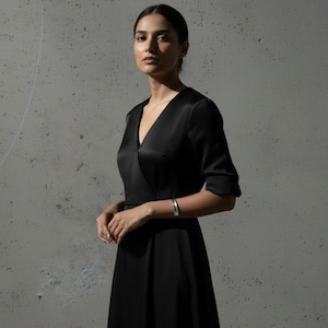 Model in a black satin wrap dress wearing a minimalist silver cuff bracelet. The clean lines and subtle sheen of the cuff echo the simplicity of the outfit. Shot against a concrete wall, the image balances elegance and raw texture with calm, composed lighting.
