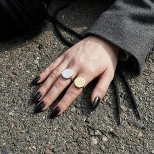 hand with black pointed nails wearing two round signet rings in 925 silver and 14k gold, resting on rough asphalt surface, neutral sleeve visible
