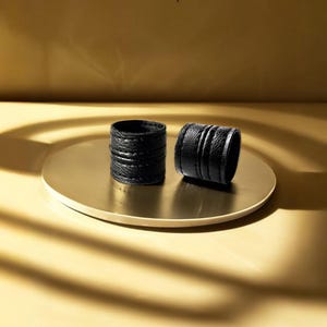 Two black vegan leather wrap rings displayed on a round brass pedestal, photographed against a warm golden background with soft directional light and curved shadows. Minimal leather ring cuffs with layered band construction and visible grain texture, shown off-hand to highlight thickness, form, and material contrast. Sculptural contemporary jewelry by Rannka, styled in a refined editorial setting with warm tones and architectural composition, suitable for modern stacking and thoughtful gifting.