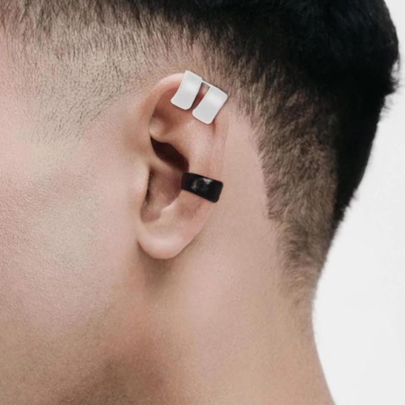 Close up side profile of a male model with short dark hair, wearing matte white and matte black ear cuffs, natural skin texture, tattoo visible on the back of the neck, photographed against a neutral background.