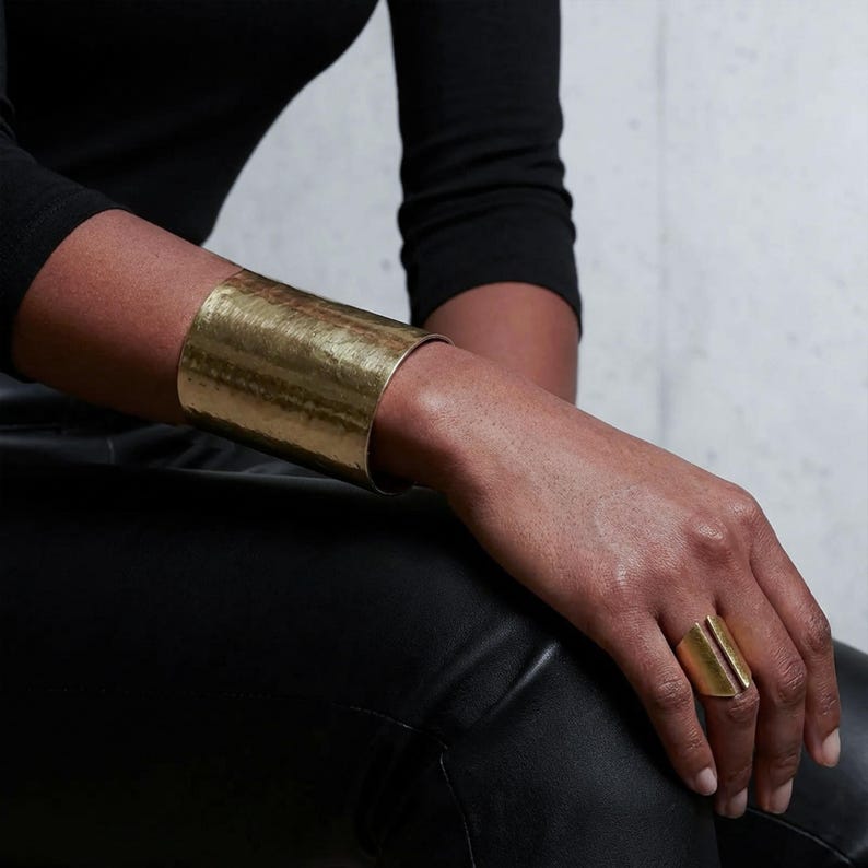 Close-up of a model’s hand resting on black leather pants, wearing a wide matte gold cuff bracelet and a sculptural open gold ring, natural skin texture visible, minimal high-end editorial styling, soft diffused studio lighting, modern architectural jewelry, refined luxury mood, Rannka jewelry.