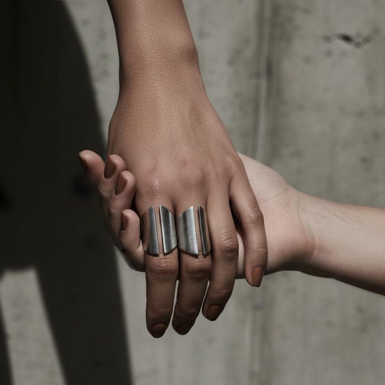 Close-up of two hands gently intertwined, one wearing two sculptural silver statement rings with a bold open-ended design, captured in soft natural light against a minimalist concrete background.