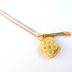 May include: A gold chain necklace with a pendant of a miniature peanut butter and banana sandwich. The sandwich is made of white bread with a brown peanut butter filling and four yellow banana slices. The pendant is attached to a small gold knife with a pink peanut butter spread.