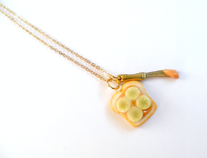 May include: A gold chain necklace with a pendant of a miniature peanut butter and banana sandwich. The sandwich is made of white bread with a brown peanut butter filling and four yellow banana slices. The pendant is attached to a small gold knife with a pink peanut butter spread.