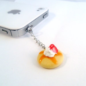 May include: A small, yellow pancake charm with a white dollop of whipped cream and two red strawberry slices on top. The charm is attached to a silver chain.