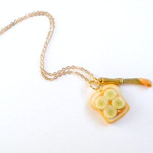 May include: A gold chain necklace with a pendant of a slice of bread with peanut butter and banana slices. The pendant is attached to a small gold knife.