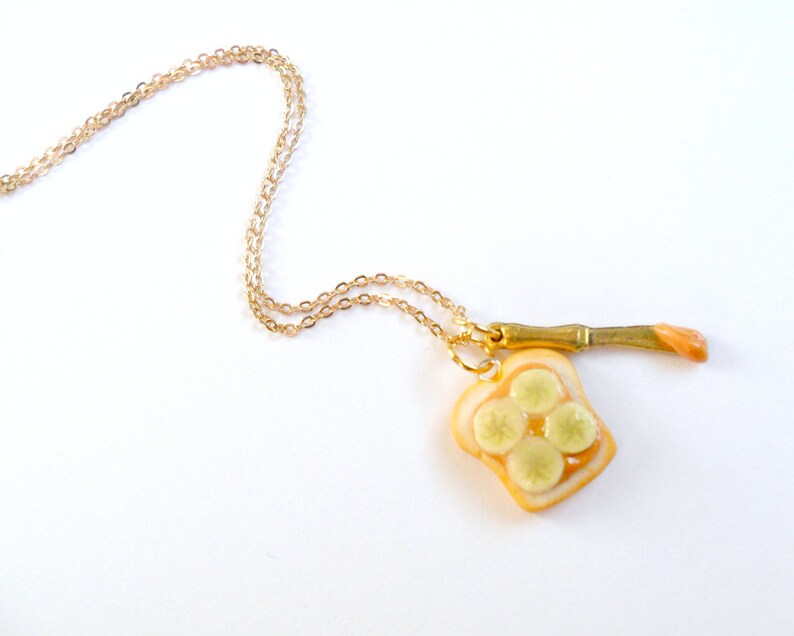 May include: A gold chain necklace with a pendant of a slice of bread with peanut butter and banana slices. The pendant is attached to a small gold knife.