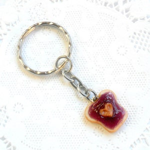 May include: A silver key chain with a small, handmade peanut butter and jelly sandwich charm. The sandwich is made of brown bread with a heart-shaped dollop of peanut butter and a layer of purple jelly.