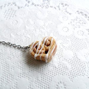 May include: A heart-shaped cinnamon roll keychain with white icing. The keychain is made of polymer clay and is attached to a silver chain.