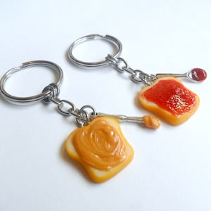 May include: A pair of keychains featuring miniature bread slices with peanut butter and jelly. The keychains are made of polymer clay and have silver key rings.
