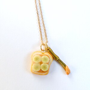 May include: A gold chain necklace with a pendant of a miniature peanut butter and jelly sandwich and a miniature knife. The sandwich is made of two slices of bread with peanut butter and jelly filling.