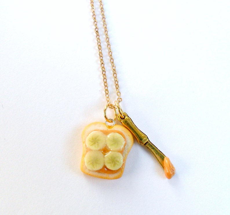 May include: A gold chain necklace with a pendant of a miniature peanut butter and jelly sandwich and a miniature knife. The sandwich is made of two slices of bread with peanut butter and jelly filling.