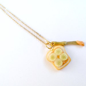 May include: A gold chain necklace with a pendant of a slice of bread with peanut butter and banana slices. The pendant is attached to a small gold knife.