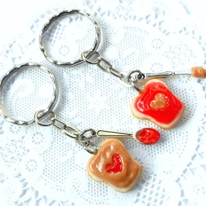 May include: Two silver keychains with a peanut butter and jelly sandwich charm. The charms are made of polymer clay and feature a heart-shaped piece of jelly on top of a peanut butter spread. One charm has a red jelly and the other has a brown peanut butter spread.