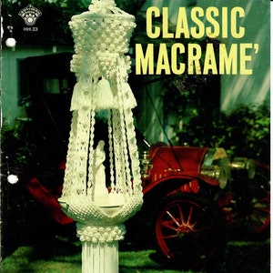 May include: A white macrame hanging with a white ceramic figurine inside. The macrame is made with a variety of knots and has a tassel at the bottom. The hanging is in front of a red car and green grass.