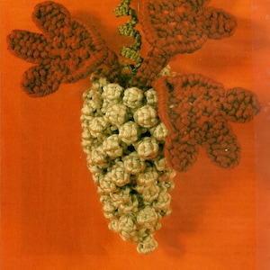 Op de afbeelding: Een gehaakte wijnstok met groene bladeren en een tros beige druiven. De druiven zijn gemaakt van garen en zijn in een tros gerangschikt.