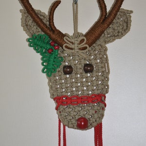 May include: A brown and beige crocheted reindeer head with red and green accents. The reindeer has a red nose, brown eyes, and a green holly sprig on the side of its head. The reindeer is hanging from a white string.