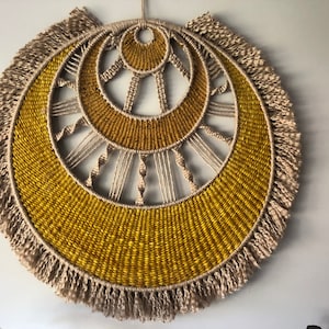May include: A large, round wall hanging made of woven natural fibers. The center of the hanging features a concentric circle design in shades of yellow and beige. The outer edge of the hanging is trimmed with a fringe of natural fibers.