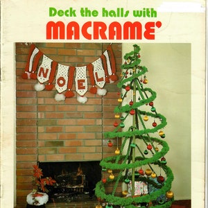May include: A green macrame Christmas tree with red and gold ornaments, decorated with a white and red crocheted "NOEL" banner. The tree is in front of a brick fireplace with a lit fire.