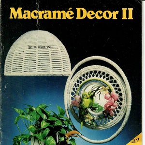 Peut inclure: Une couverture de livre pour un livre de projets de décoration en macramé intitulé "Macramé Decor II" présentant un support de plante suspendu en macramé blanc avec deux oiseaux verts et une lampe suspendue en macramé blanc. La couverture du livre est jaune et porte le texte "Featuring Decorator Plant Stands".