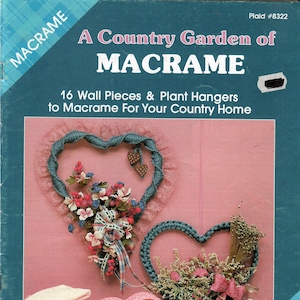 May include: A book cover with a pink background featuring a macrame heart-shaped wall hanging with flowers and a macrame plant hanger with dried flowers. The book title is "A Country Garden of Macrame" with the subtitle "16 Wall Pieces & Plant Hangers to Macrame For Your Country Home". The book is by Sandy Dye.