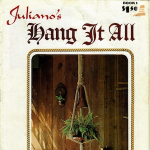 Può includere: Una copertina di libro con il titolo "Juliano's Hang It All" in lettere rosse. La copertina presenta una foto di un portavasi in macramè con una pianta di felce appesa. Il portavasi è fatto di corda naturale e ha una base in legno. La foto è scattata contro un muro di legno con una lanterna e una botte di legno sullo sfondo. Il testo "Baba's Lamp" è scritto sotto la foto.