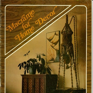 Può includere: Una copertina di libro con il titolo "Macramé per l'arredamento della casa" in un carattere marrone e arancione. La copertina presenta un porta piante sospeso in macramé con una corda marrone e una pianta verde. Il libro fa parte di una serie chiamata "This is a Handy Craft Pak Book".
