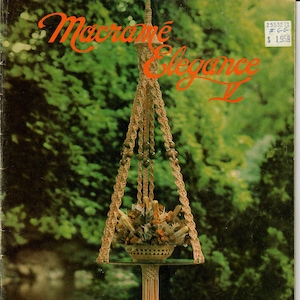 May include: A macrame plant hanger with a brown and beige cord, hanging from a ceiling. The hanger has a round, brown pot with dried flowers inside. The text "Macrame Elegance II" is in the top left corner of the image. The text "Belle Pensée" is in the bottom right corner of the image.