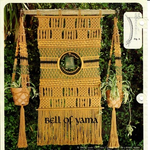 以下が含まれることがあります： 中央にベルがついたマクラメの壁掛けで、タイトルは「ベル・オブ・ヤマ」です。壁掛けは茶色、緑、黄色の紐で作られており、上部に木製の棒があります。