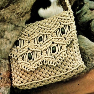 Op de afbeelding: Een beige macramé tas met een geweven patroon en zwarte kralen. De tas heeft een lange band en wordt over de schouder gedragen.