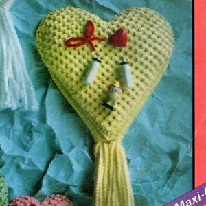 May include: A yellow heart-shaped crocheted ornament with a red bow, a red heart, and a small doll figure. The ornament has a long yellow tassel. The text "Made with Maxi-Craft Twisted Hercu" is visible in the image.