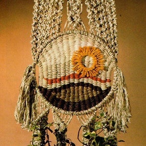 May include: A macrame plant hanger with two pots and green plants. The hanger is made of natural beige cord and has a circular design at the top. The pots are brown and the plants are green.