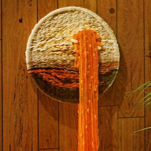May include: A round woven wall hanging with a brown, orange, and white design. A long orange yarn tassel hangs from the bottom.