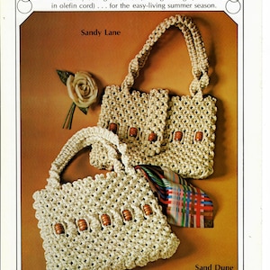 以下が含まれることがあります： ナチュラルカラーのコードで編まれた2つの手編みバッグ。大きなバッグは長いストラップが付いており、木製ビーズで飾られています。小さなバッグは短いストラップが付いており、木製ビーズで飾られています。バッグは、「Noteworthy Neutrals」、「Sandy Lane」、「Sand Dune」というテキストが書かれた茶色の背景に飾られています。