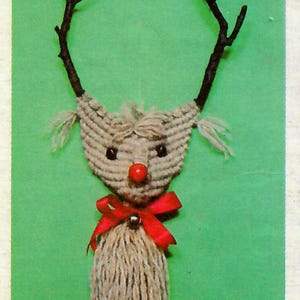 May include: A handmade reindeer ornament made from yarn and twigs. The reindeer has a red nose, a red bow, and a bell. The ornament is hanging on a green background.