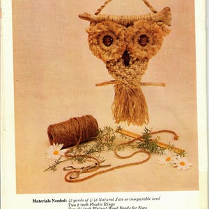 Op de afbeelding: Een handgemaakte macramé uil wanddecoratie met een natuurlijke jute textuur. De uil heeft twee grote cirkelvormige ogen, een snavel en een decoratieve franje. De afbeelding toont ook benodigde materialen: jute, plastic ringen, houten kralen, drijfhout, schaar en bloemen.