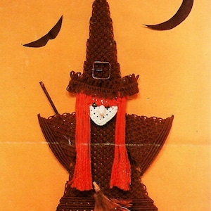 May include: A brown macrame wall hanging shaped like a witch, featuring a tall pointed hat with a buckle, long bright orange yarn hair, a white triangular face, a small broomstick, and two small brown shoes with buckles at the bottom.