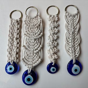 May include: Four handmade macrame keychains in varying designs. Each keychain is off-white with a gold-colored ring and a blue glass eye charm. The macrame patterns include woven, knotted, and braided styles.
