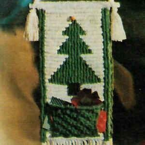 May include: A white yarn wall hanging with a green Christmas tree design. The tree is surrounded by green and red yarn ornaments. The hanging has a fringe border.