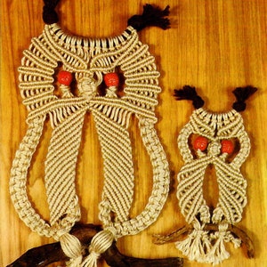 May include: Two macrame owls, one large and one small, are hanging from a wooden branch. The owls are made with natural colored string and have red bead eyes.